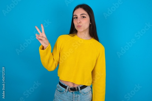 young brunette girl wearing yellow knitted sweater against blue wall makes peace gesture keeps lips folded shows v sign. Body language concept
