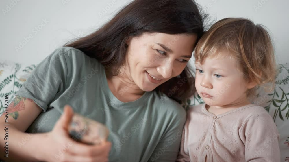 Asian young mother and curly boy daughter toddler kid look at phone screen video chat online. Woman and baby taking selfie or watch cartoons. Spend time together. Parenthood concept. bedroom morning Asian young mother and curly boy daughter toddler kid look at phone screen video chat online. Woman and baby taking selfie or watch cartoons. Spend time together. Parenthood concept. bedroom morning