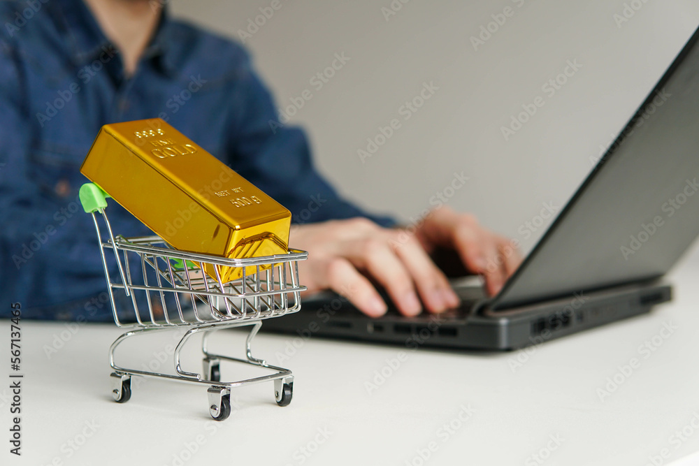 Small shopping cart with gold bar on the table. Man using laptop for online trading or buying gold bars for investment.