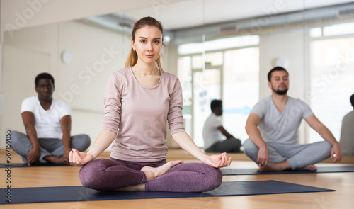 Young woman sitting in lotus pose on mat during group yoga training.