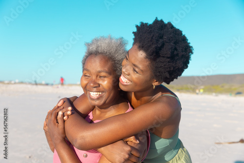 Cheerful african american young woman embracing senior mother and enjoying at beach against blue sky