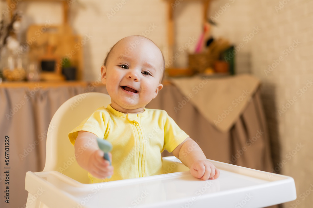a cute baby in a yellow bodysuit is sitting in a high chair in the kitchen of the house with a plastic spoon. Baby food, first complementary food a cute baby in a yellow bodysuit is sitting in a high chair in the kitchen of the house with a plastic spoon. Baby food, first complementary food
