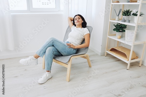 Woman sitting in a chair listening to music on wireless headphones at home in jeans and a white T-shirt, fall lifestyle