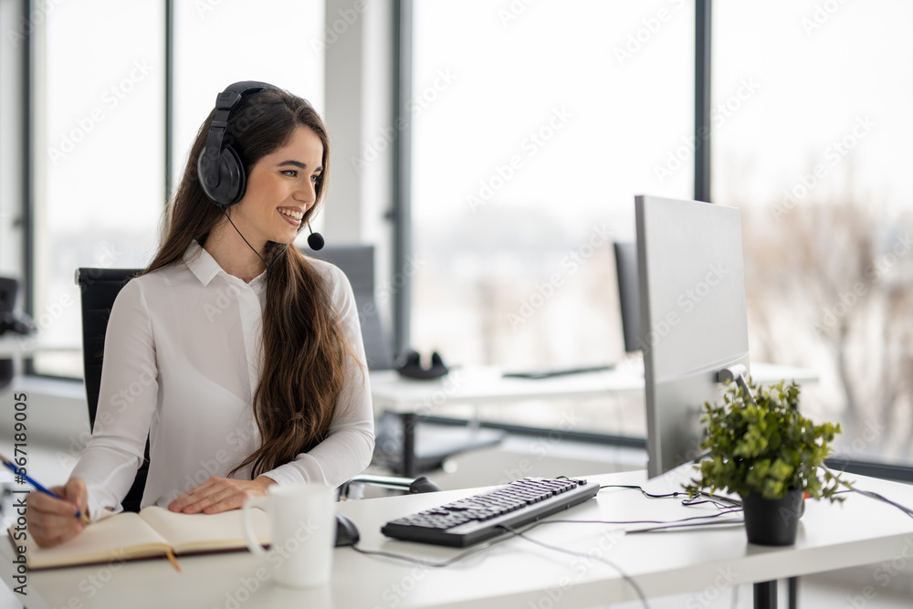 Beautiful young customer support female operator talking with client via hands-free headphones with microphone and writing notes to notebook in bright office. Beautiful young customer support female operator talking with client via hands-free headphones with microphone and writing notes to notebook in bright office.