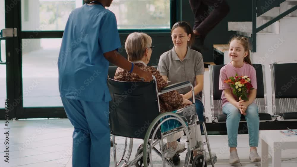 Elderly patient in wheelchair meeting with family in hospital waiting room, talking to woman and little girl in lobby. Senior person with disability receiving medical assistance at health center.