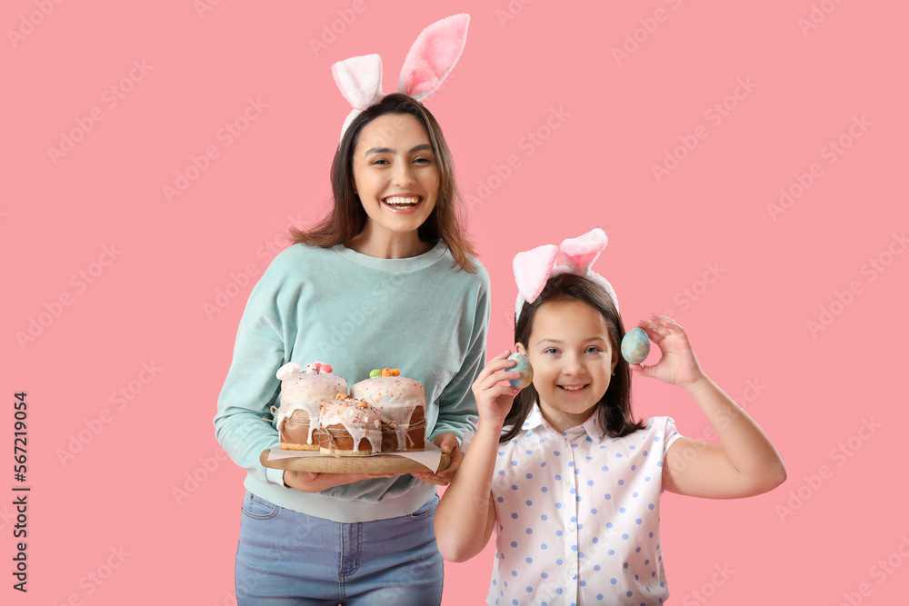 Beautiful woman and her little daughter in bunny ears with Easter cakes and eggs on pink background Beautiful woman and her little daughter in bunny ears with Easter cakes and eggs on pink background