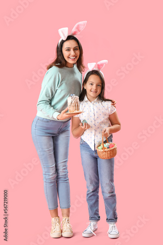 Beautiful woman and her little daughter in bunny ears with Easter cake and eggs on pink background