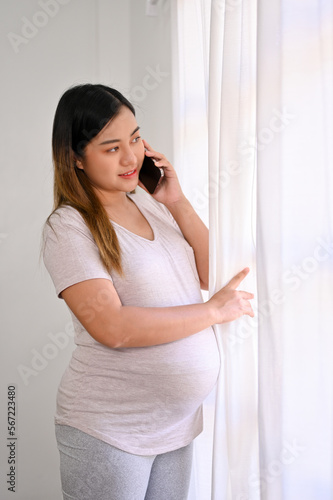 Happy Asian pregnant woman looking out the window and talking on the phone with her husband. © bongkarn