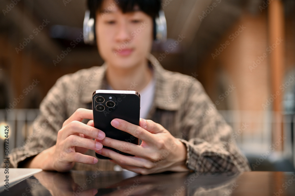 Handsome young Asian man using his smartphone while listening to music through his headphones Handsome young Asian man using his smartphone while listening to music through his headphones