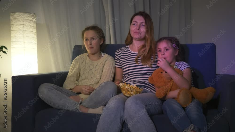 Happy family mom with daughters watching TV on the couch at night enjoying popcorn. Happy family concept, little daughters with mom watching tv program while sitting on sofa at home Happy family mom with daughters watching TV on the couch at night enjoying popcorn. Happy family concept, little daughters with mom watching tv program while sitting on sofa at home