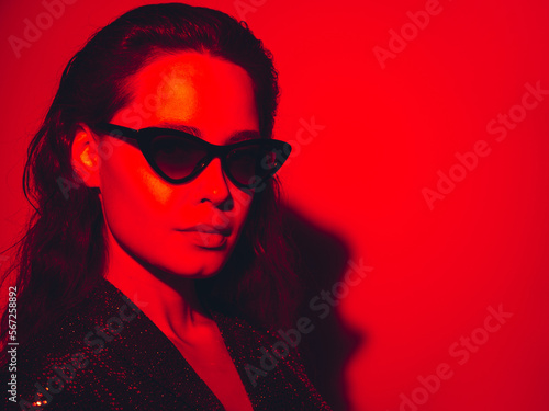 Young beautiful confident brunette female in trendy evening shiny green reflecting dress. Sexy carefree woman isolated on red neon light in studio. Fashionable model in sunglasses. high fashion