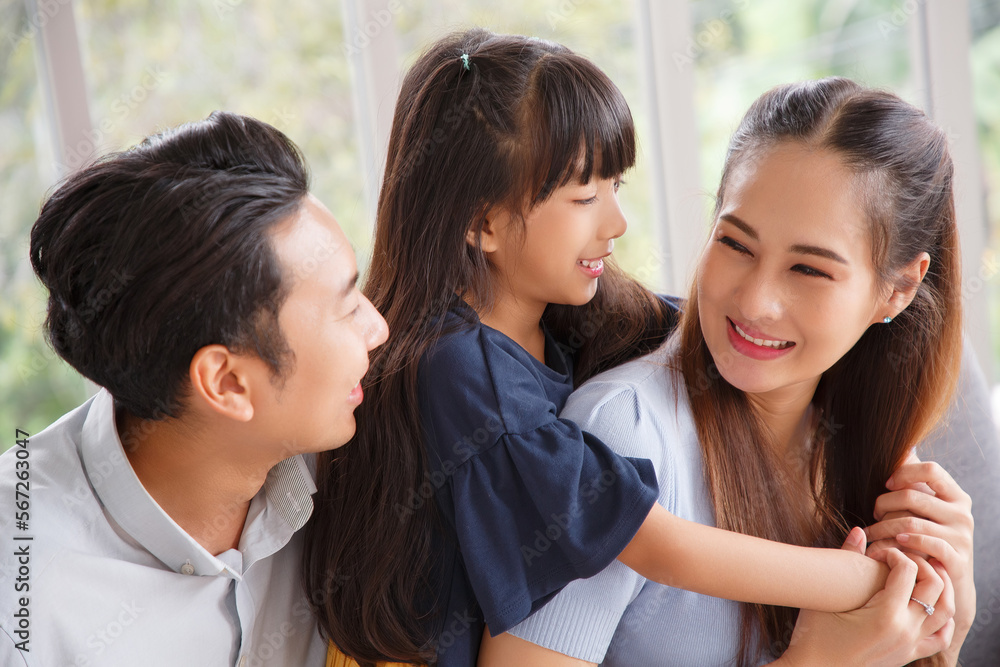 Portrait of a happy young family. Mom, dad and daughter look at the camera and smile sitting on sofa in living room. The faces of Asian parents and their child.
