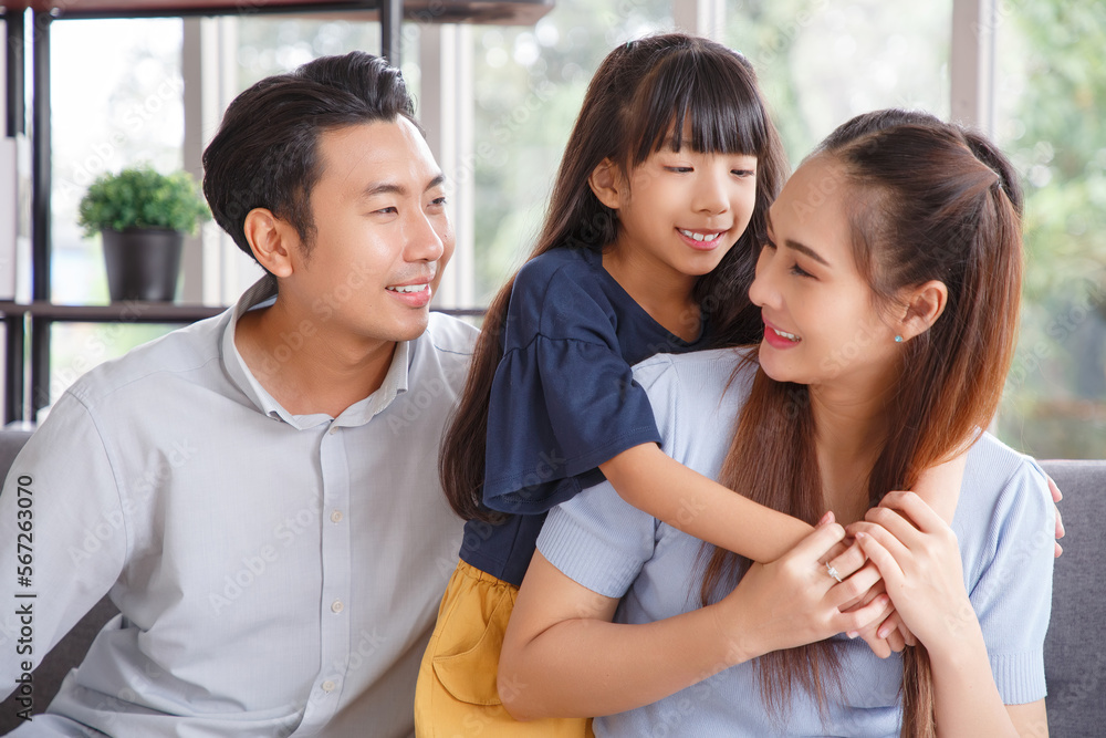 Portrait of a happy young family. Mom, dad and daughter look at the camera and smile sitting on sofa in living room. The faces of Asian parents and their child.