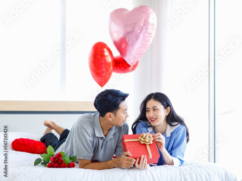 Asian young romantic cheerful lover couple male boyfriend laying down on bed giving present wrapped gift box surprise female girlfriend in bedroom decorate with pink heart shape balloon on valentine