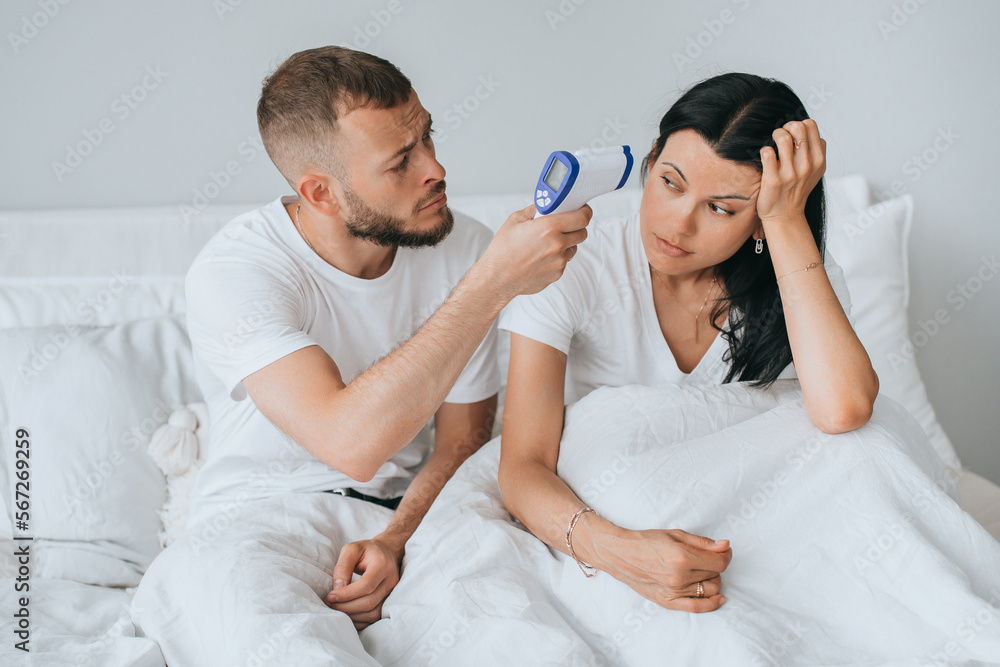 Perplexed young beardy man in bed with wife measuring temperature of woman. Ill woman at home. Caucasian guy takes care of girlfriend waiting for doctor. Medicine, virus, treatment. Health care. Perplexed young beardy man in bed with wife measuring temperature of woman. Ill woman at home. Caucasian guy takes care of girlfriend waiting for doctor. Medicine, virus, treatment. Health care.