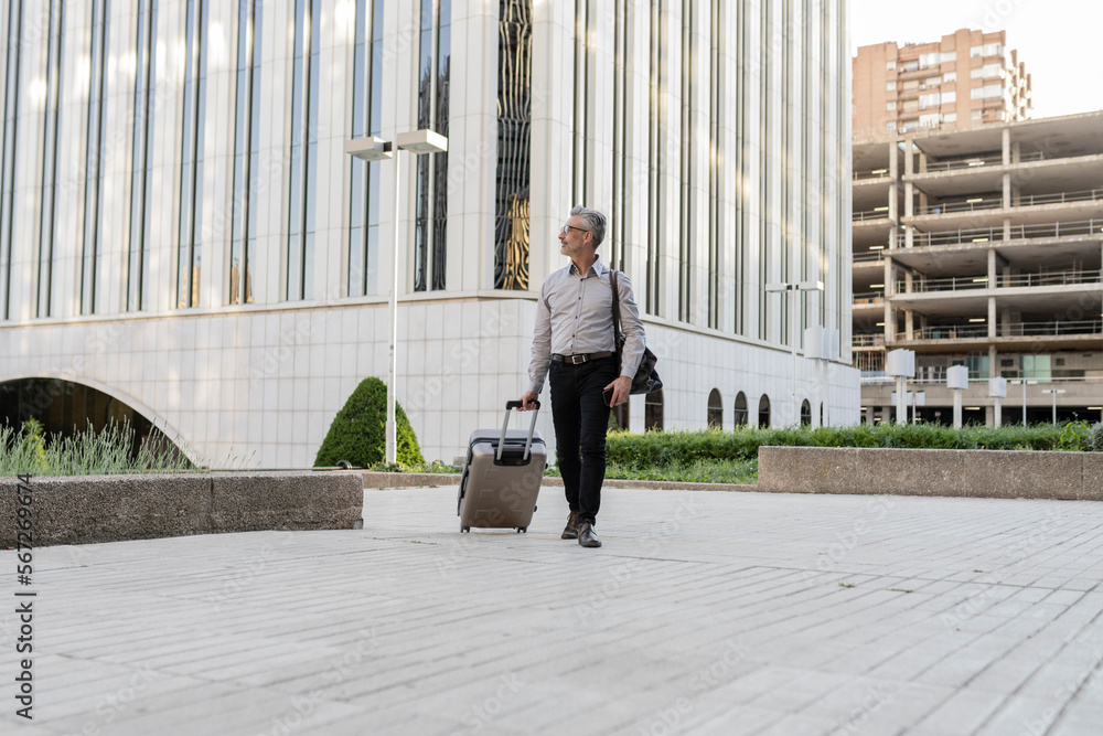 Senior businessman with suitcase, male professionals in suit walking on sidewalk next to business office building starting business trip.