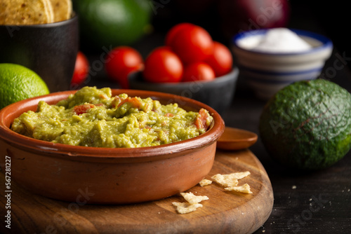 Close-up of bowl with guacamole on dark table with tomatoes, avocados, limes and salt, selective focus, horizontal