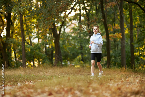 a caucasian girl in a blue sweatshirt puts on headphones in the park and prepares to run. pretty young woman puts on headphones. Pretty smiling woman puts on headphones in the park before jogging.