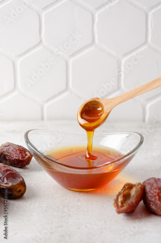Natural organic healthy date syrup flows from wood spoon into a glass bowl with whole dates on the background. Copy space