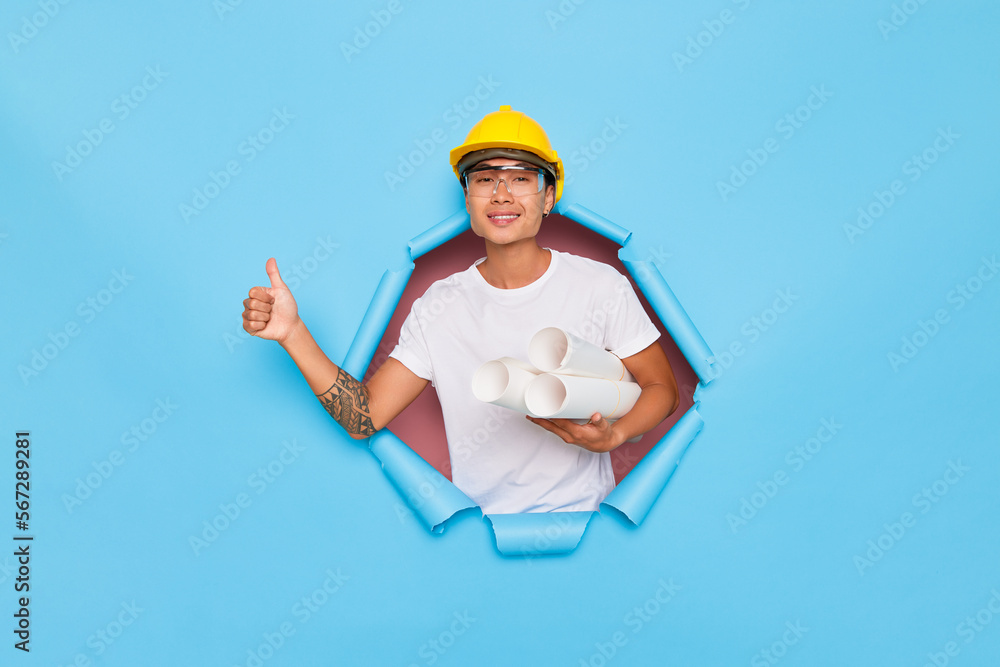 Young asian engineer in yellow helmet holds white roll paper projects smiles and gesturing thumbs up in blue background torn Young asian engineer in yellow helmet holds white roll paper projects smiles and gesturing thumbs up in blue background torn