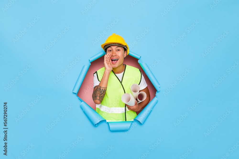 Young asian engineer Announcing somthing to the audiance wearing a yellow helmet holds a bunch of paper roll projects, screams to advertisement copy place, isolated next to torn blue background Young asian engineer Announcing somthing to the audiance wearing a yellow helmet holds a bunch of paper roll projects, screams to advertisement copy place, isolated next to torn blue background