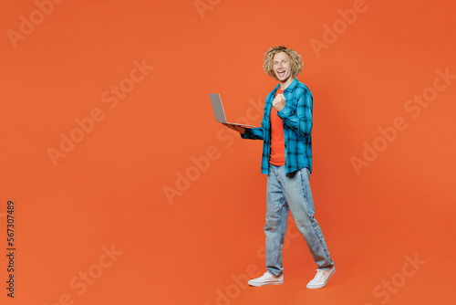 Full body young blond caucasian IT man wear blue shirt orange t-shirt hold use work on laptop pc computer do winner gesture isolated on plain red background studio portrait. People lifestyle concept.