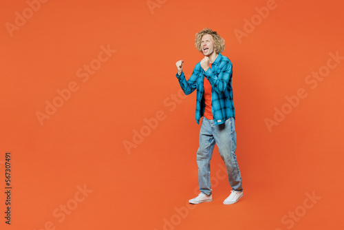 Full body fun young blond caucasian man in blue shirt orange t-shirt doing winner gesture celebrate clenching fists say yes isolated on plain red background studio portrait. People lifestyle concept
