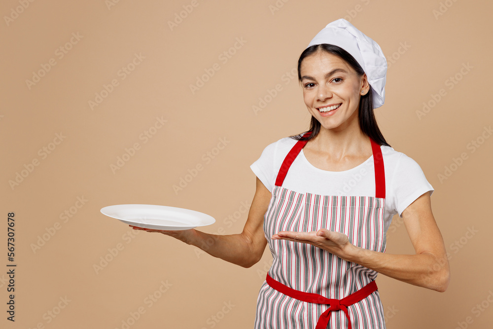 Young smiling happy housewife housekeeper chef baker latin woman wear striped apron toque hat hold in hand point hand on empty plate isolated on plain pastel light beige background. Cook food concept. Young smiling happy housewife housekeeper chef baker latin woman wear striped apron toque hat hold in hand point hand on empty plate isolated on plain pastel light beige background. Cook food concept.