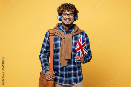 Young teen Indian boy IT student he wear casual clothes shirt glasses bag headphones listen to music hold British flag isolated on plain yellow color background High school university college concept