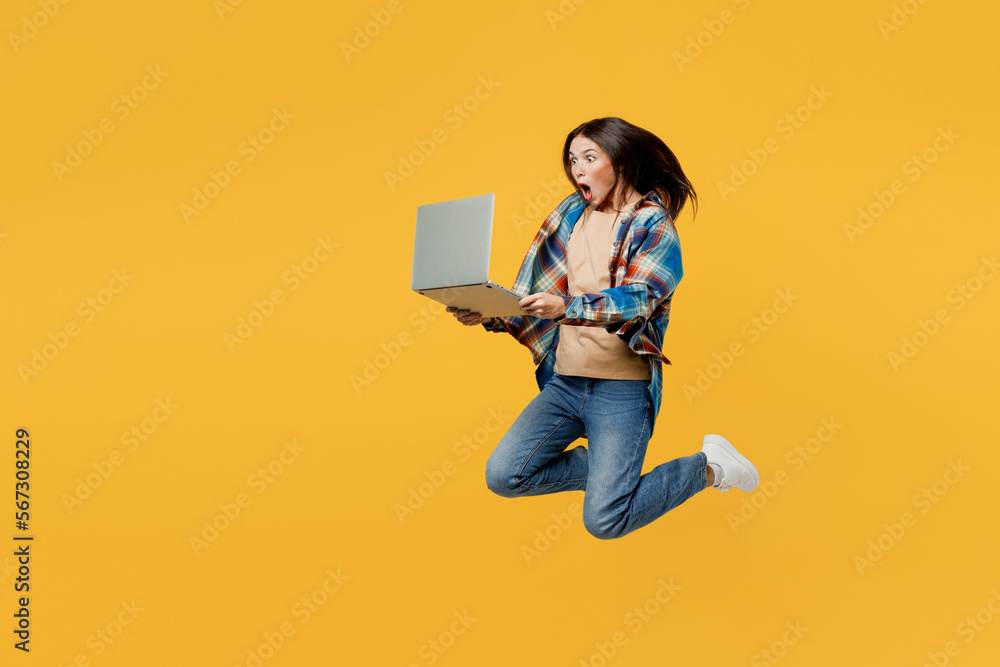 Full body side view young smiling IT woman wearing blue shirt beige t-shirt jump high hold use work on laptop pc computer isolated on plain yellow background studio portrait. People lifestyle concept. Full body side view young smiling IT woman wearing blue shirt beige t-shirt jump high hold use work on laptop pc computer isolated on plain yellow background studio portrait. People lifestyle concept.