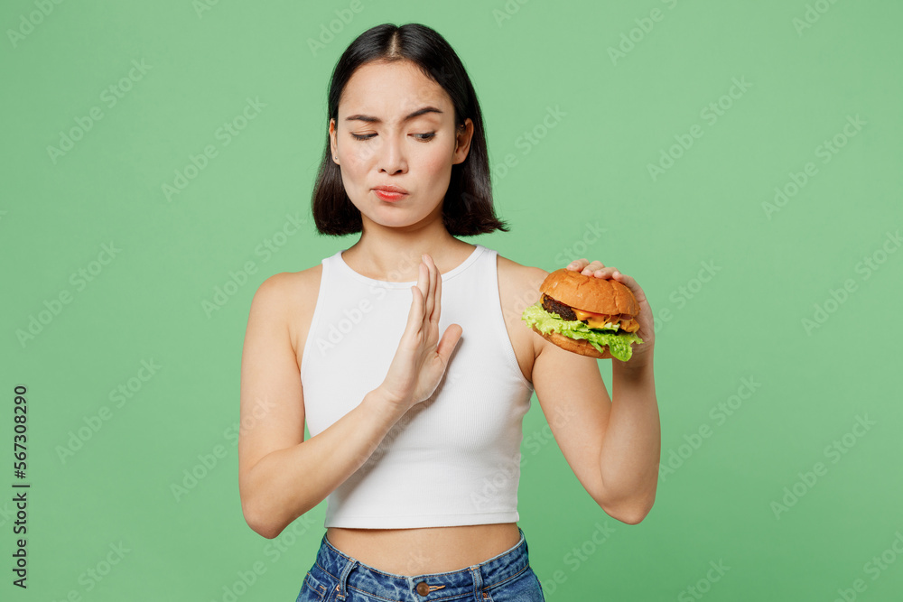 Young sad woman wear white clothes hold eat burger show hand stop gesture say no isolated on plain pastel light green background. Proper nutrition healthy lifestyle fast food unhealthy choice concept. Young sad woman wear white clothes hold eat burger show hand stop gesture say no isolated on plain pastel light green background. Proper nutrition healthy lifestyle fast food unhealthy choice concept.