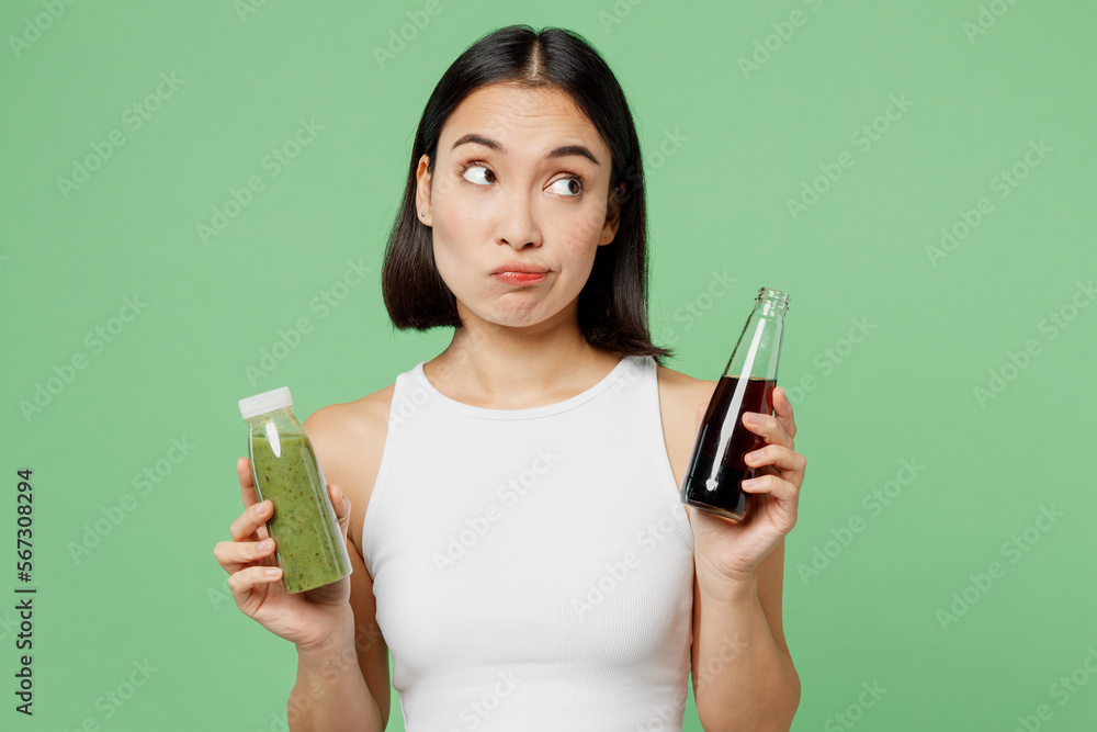 Young woman wear white clothes hold pressed juice vegetable smoothie soda pop look aside isolated on plain pastel light green background. Proper nutrition healthy fast food unhealthy choice concept. Young woman wear white clothes hold pressed juice vegetable smoothie soda pop look aside isolated on plain pastel light green background. Proper nutrition healthy fast food unhealthy choice concept.