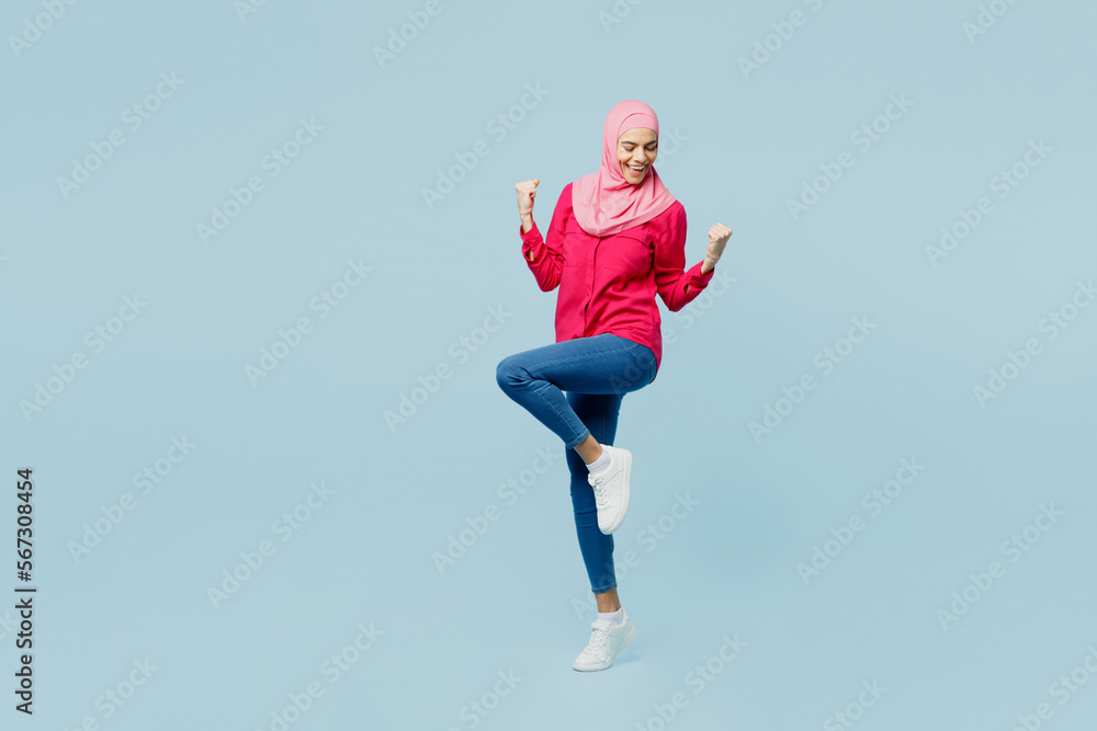 Full body young arabian asian muslim woman wear pink abaya hijab do winner gesture clench fist isolated on plain pastel light blue cyan background studio portrait. People uae islam religious concept.