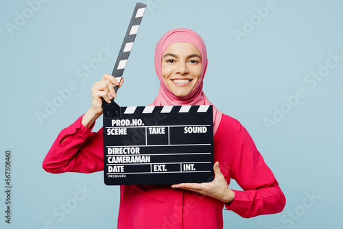 Young arabian muslim woman wearing pink abaya hijab hold in hand classic black film making clapperboard isolated on plain pastel light blue cyan background studio. People uae islam religious concept. © ViDi Studio