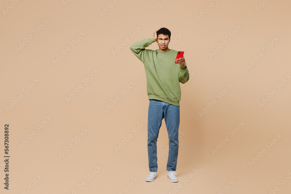 Fototapeta premium Full body sad young man of African American ethnicity wear green sweatshirt hold scratch head use mobile cell phone isolated on plain pastel light beige background studio . People lifestyle concept.