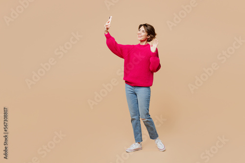 Full body young woman wear pink sweater doing selfie shot on mobile cell phone post photo on social network isolated on plain pastel light beige background studio portrait. People lifestyle concept.