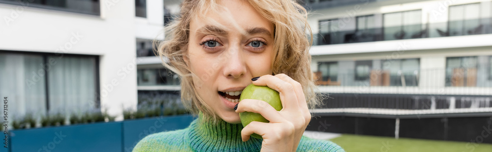 blonde woman in sweater eating green apple near hotel building, banner. blonde woman in sweater eating green apple near hotel building, banner.