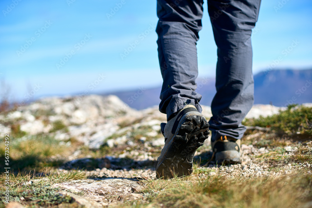 Legs of a hiker in trekking boots walking in the mountains closeup shot. Feet of walking tourist wearing trekking shoes on a rocky road captured from behind. Hiking male wearing pants and boots walk Legs of a hiker in trekking boots walking in the mountains closeup shot. Feet of walking tourist wearing trekking shoes on a rocky road captured from behind. Hiking male wearing pants and boots walk