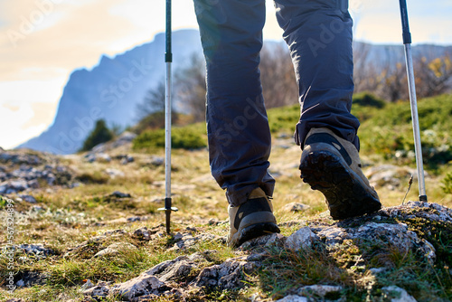Legs of a hiker in trekking boots walking in the mountains with Nordic walking poles closeup shot. Feet of walking tourist wearing trekking shoes with hiking sticks on rocky road captured from behind