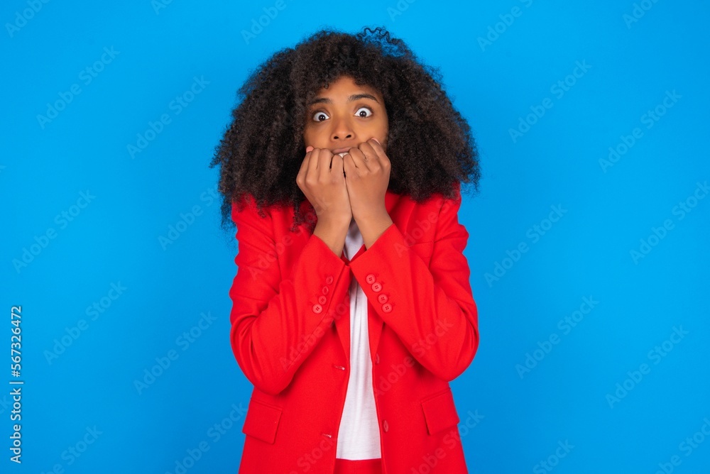 Anxiety - young businesswoman with afro hairstyle wearing red over blue background covering his mouth with hands scared from something or someone bitting nails. Anxiety - young businesswoman with afro hairstyle wearing red over blue background covering his mouth with hands scared from something or someone bitting nails.