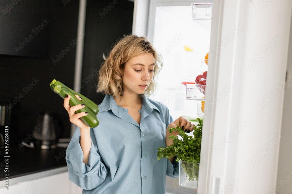 blonde young woman in blue shirt looking at greenery while holding zucchini near open refrigerator in kitchen. blonde young woman in blue shirt looking at greenery while holding zucchini near open refrigerator in kitchen.