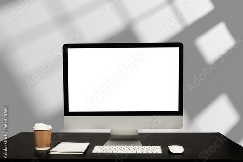 Computer monitor with white blank screen putting on white working desk with wireless mouse and keyboard over blurred modern office as background..
