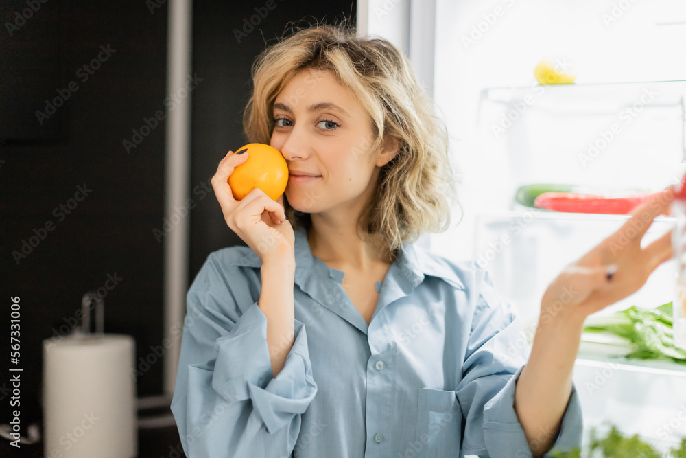 happy young woman smelling orange near refrigerator in kitchen.