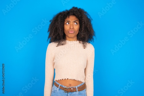 Displeased Young woman with afro hair style wearing crop top over blue background frowns face feels unhappy has some problems. Negative emotions and feelings concept photo