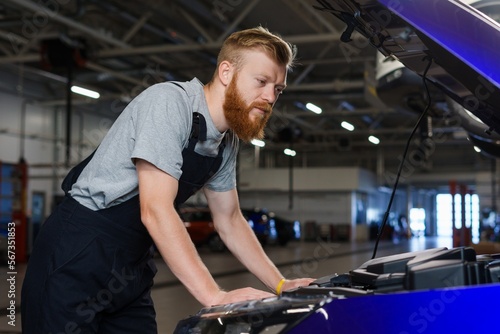 A brutal bearded man in uniform works at a car repair station. Checking the car in a clean modern space
