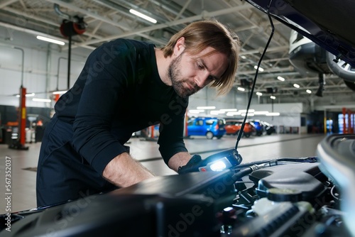 A male mechanic in uniform with a flashlight performs diagnostics under the hood of the car. Modern and technological car service