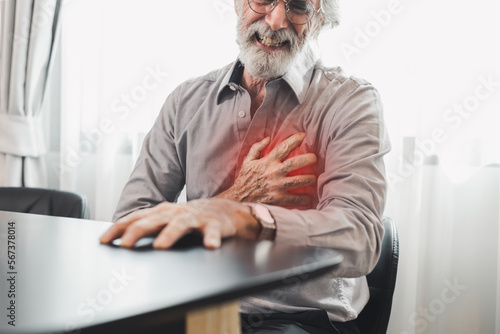 Senior man in spectacles presses hand to chest has heart attack suffers from unbearable chest pain closes eyes wears glasses sitting on the chair in living room. People age and problems with health.