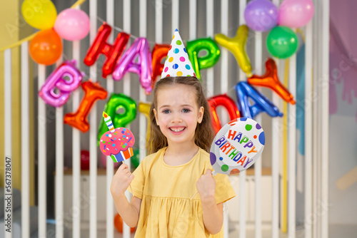 The birthday girl in a festive hat, with a pipe and props is having fun on the background of a multi-colored wall with balloons.