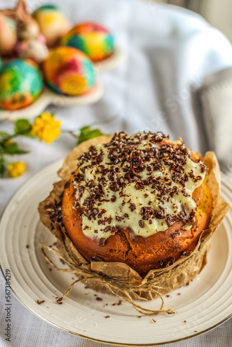 Traditional Russian Easter cake - kulich decorated with royal icing and chocolate flakes on white tablecloth background with colored eggs. Tasty handmade Orthodox paska.