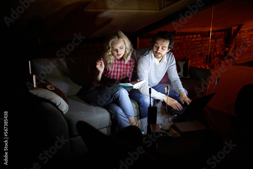 Man and woman, couple sitting on sofa at home in the evening and studying, working, reading book with little light of battery lamp. No electricity, blackout. Concept of power outage